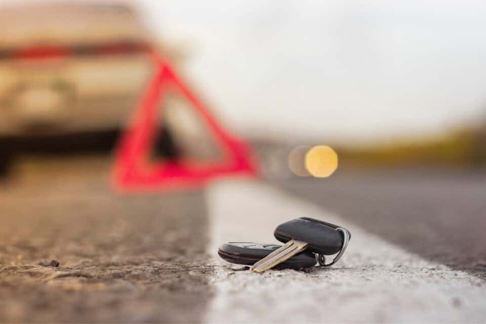 Car keys lying on a road with a blurry car and a red warning triangle in the background, as if awaiting roadside assistance.