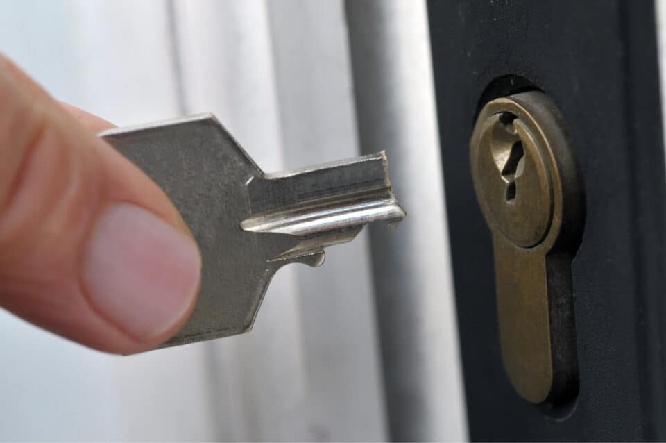 A close-up of a hand holding a broken key near a door lock, as if in need of an emergency locksmith. The key appears to have snapped, with only a small portion remaining between the fingers. The cylinder lock is mounted in the door.