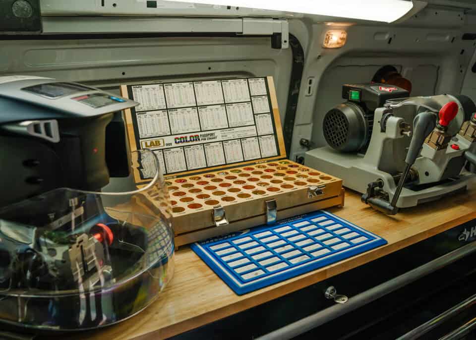 A locksmith workspace inside a van features a key-cutting machine, a color-coded layout of keys, a key identification chart, and a shielded grinder.