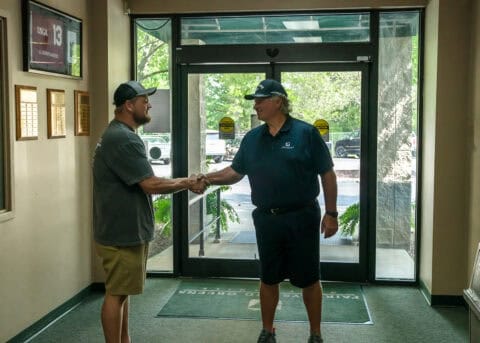Two men wearing casual clothing and caps are shaking hands in a building entrance lobby with windows and plaques on the wall, discussing key cutting services.