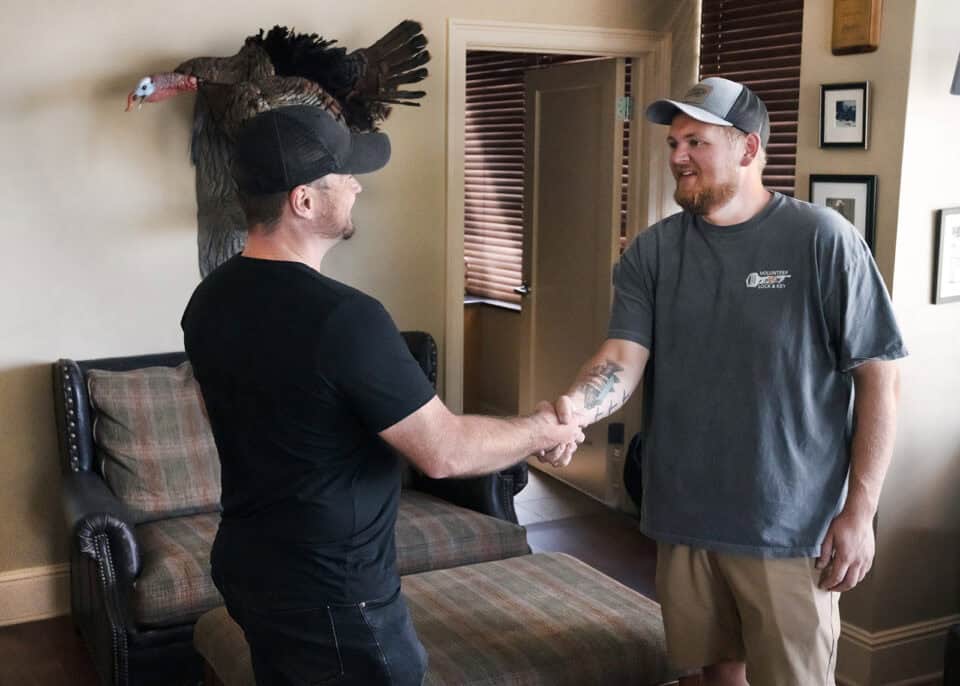 Two men in casual clothing shake hands in a room with a beige wall, wooden blinds, and a mounted turkey on the wall. One man wears a black shirt and cap, while the other wears a gray shirt and cap. The friendly interaction suggests they might both be locksmiths discussing key cutting or roadside assistance services.