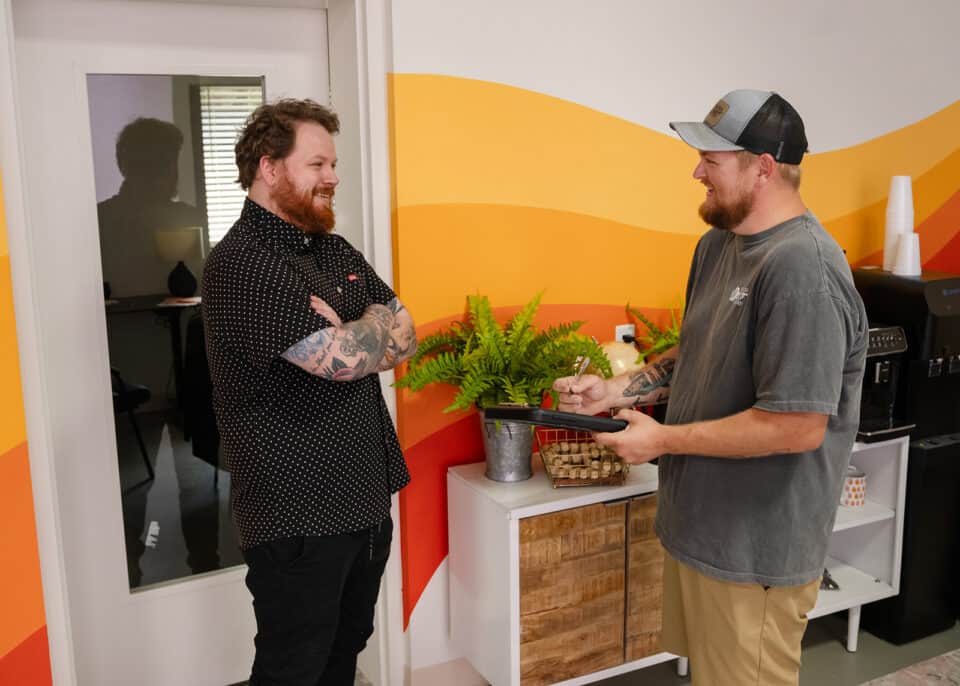 Two men are standing and talking in a brightly decorated room with a yellow and orange wall. One man, a residential locksmith, is holding a tablet while the other has his arms crossed and smiles.