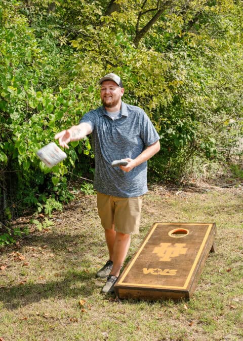 A man wearing a cap and blue shirt plays cornhole outdoors, throwing a bean bag towards a wooden board with a target hole. Trees and greenery are in the background, creating an idyllic setting that even a residential locksmith would enjoy during their downtime.