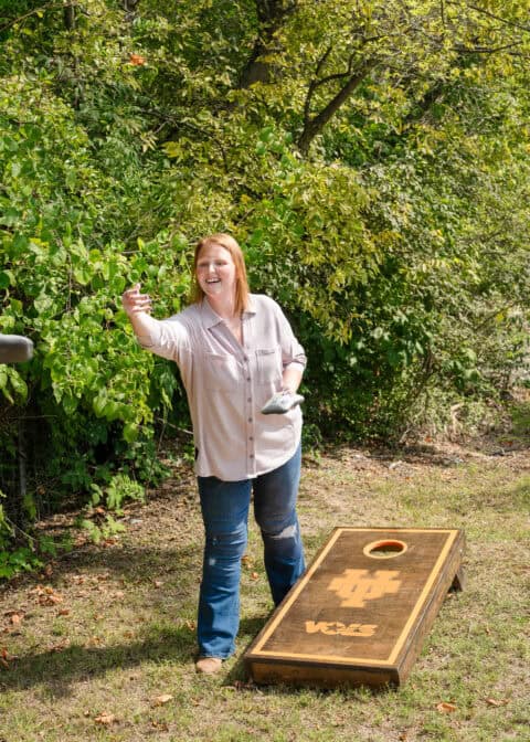 A woman is playing cornhole outside, standing next to a cornhole board and about to toss a bean bag. Amidst the trees and greenery in the background, it’s a serene setting reminiscent of peaceful moments between key cutting or lock installation appointments.