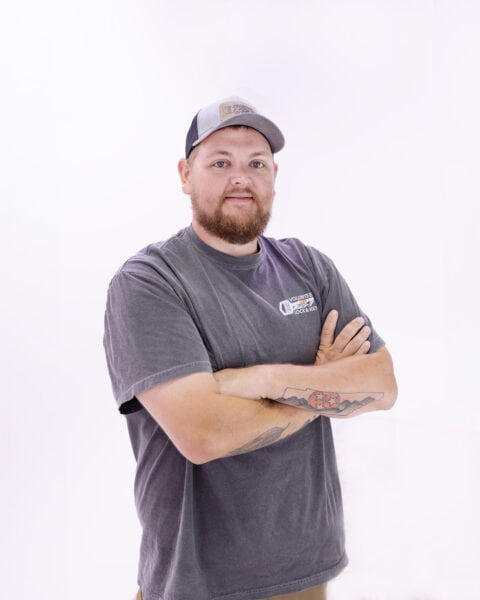 A man with a beard and tattoos wearing a cap, t-shirt, and shorts stands with arms crossed against a plain background, ready for roadside assistance.