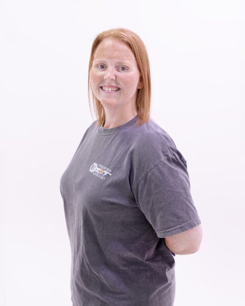 A person with straight red hair and light skin standing against a white background, wearing a dark gray T-shirt with a logo on the left side. The logo hints at their expertise as a residential locksmith.