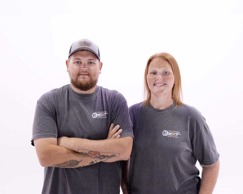 A man and a woman in matching gray t-shirts and caps with logos stand side by side against a white background. The man, possibly a locksmith, has his arms crossed while the woman, who might specialize in roadside assistance, stands with her arms at her sides.