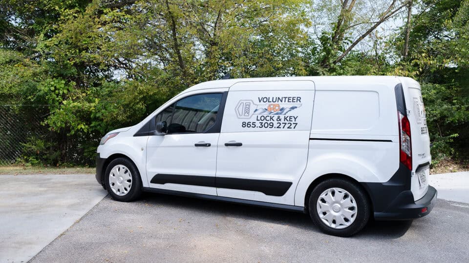 White locksmith van parked outdoors, displaying text: "Volunteer Lock & Key 865.309.2727" on the side. Trees and a chain-link fence are in the background. The van offers residential locksmith services, including key cutting and lock installation.