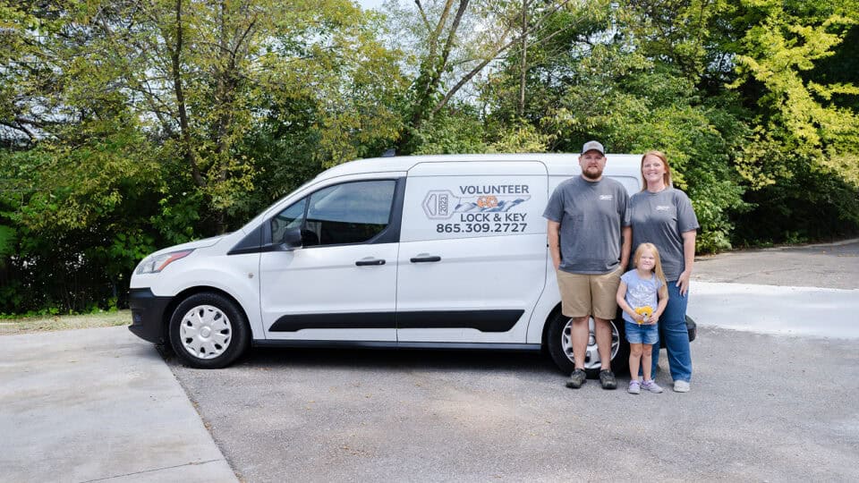 A man, woman, and child stand next to a white van labeled "Volunteer Lock & Key" offering roadside assistance in a parking lot with trees in the background.