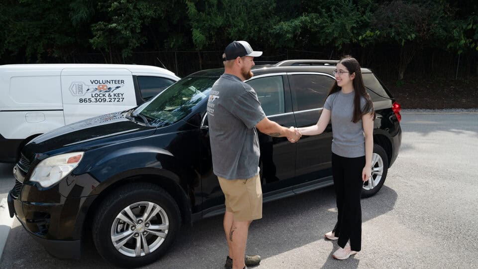 A man and a woman shake hands next to a black SUV, while a white van labeled "Volunteer Lock & Key" providing locksmith services is parked in the background.