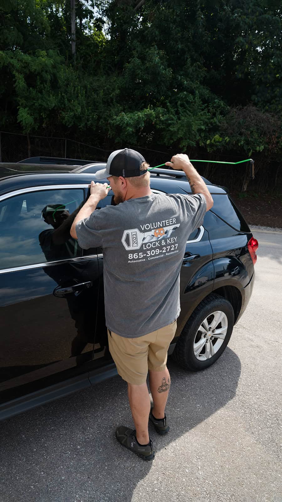 A man in a "Volunteer Lock & Key" shirt works to unlock a black car with a tool. As part of their skills in both commercial locksmith services, a green hook is inserted through the top of the car door.