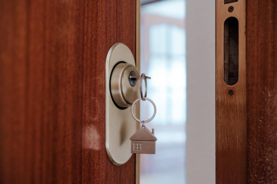 A key with a house-shaped keychain is inserted in the lock of a wooden door, which was expertly installed by a commercial locksmith. The door is partially open.